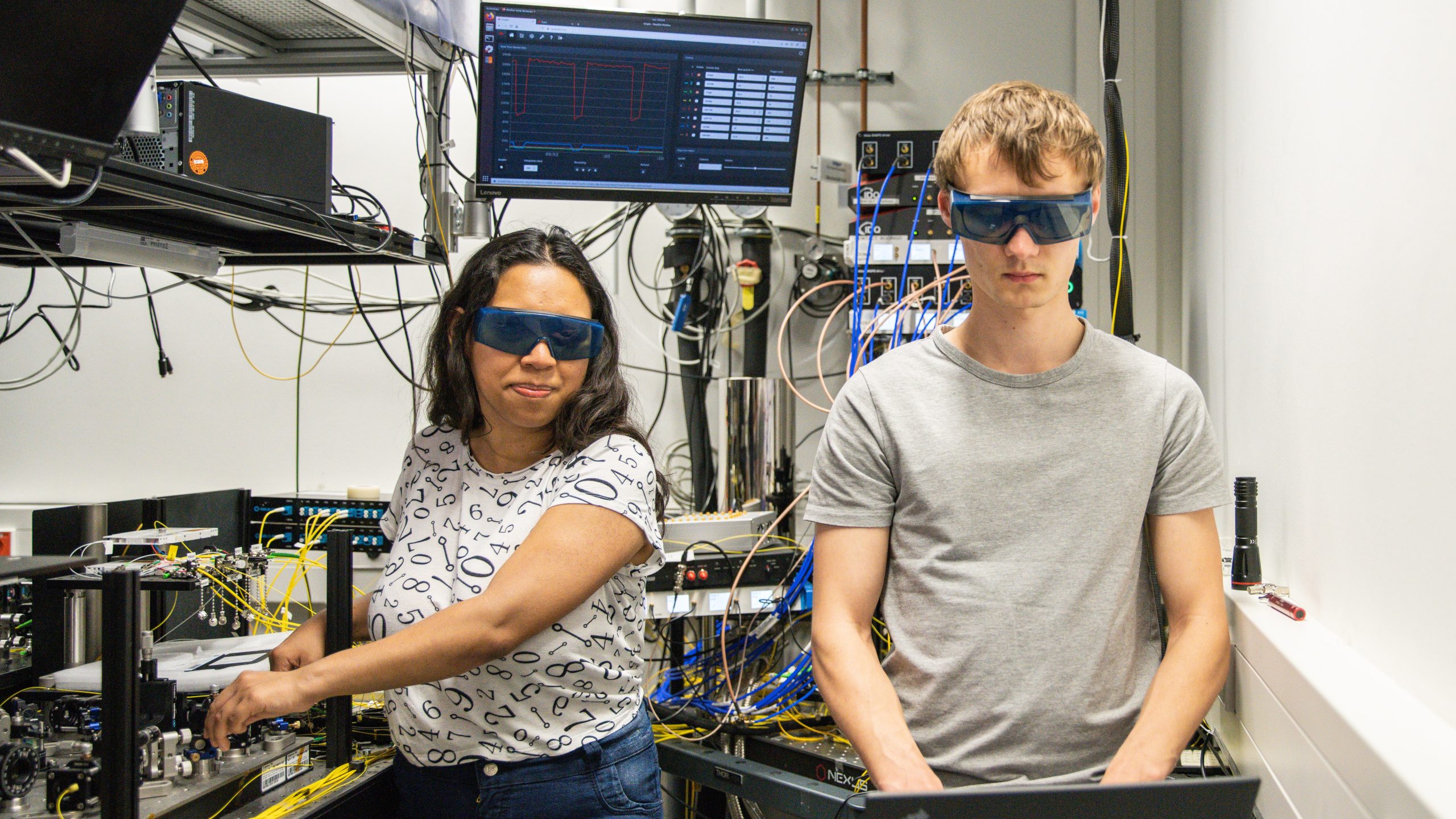 zwei Menschen stehen mit Schutzbrillen in einem Raum mit Technik. Eine Person arbeitet an der Technik und die andere Person am Laptop
